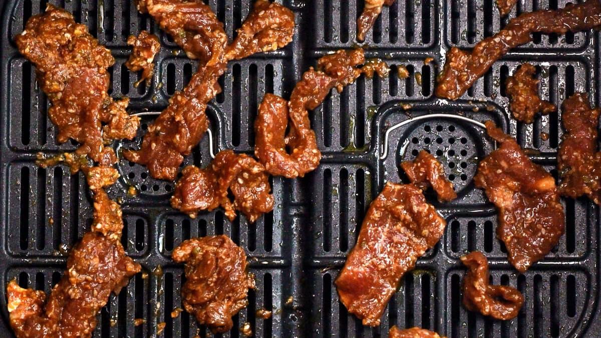 Marinated koren bbq beef strips of raw meat are spread out on a black air fryer basket, ready to be cooked. The meat is coated in a reddish-brown sauce, and the air fryer tray has a grid and perforated pattern.