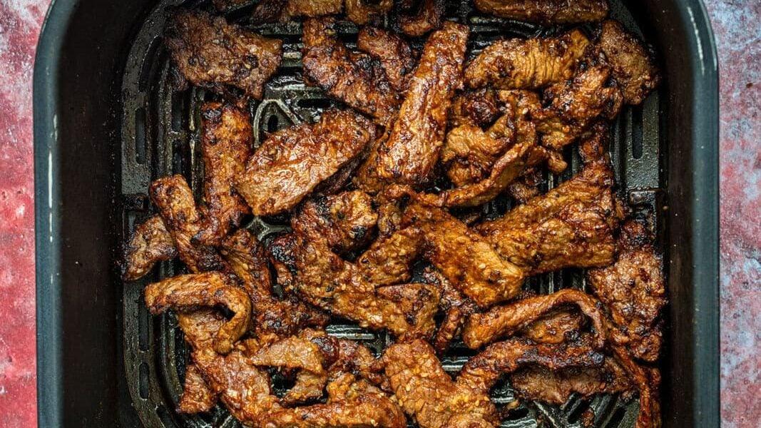 Cooked pieces of korean bbq beef in an air fryer basket, viewed from above, with a red textured background.