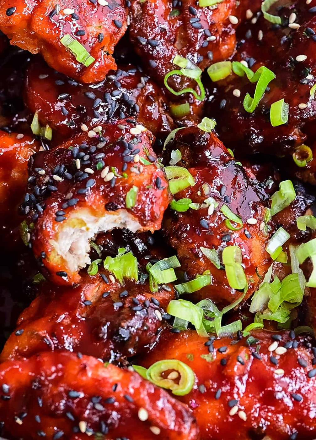 Close-up of Korean Fried Chicken topped with sliced green onions, and black sesame and white sesame seeds. The chicken is coated in a glossy, red sauce and generously garnished for extra flavor.