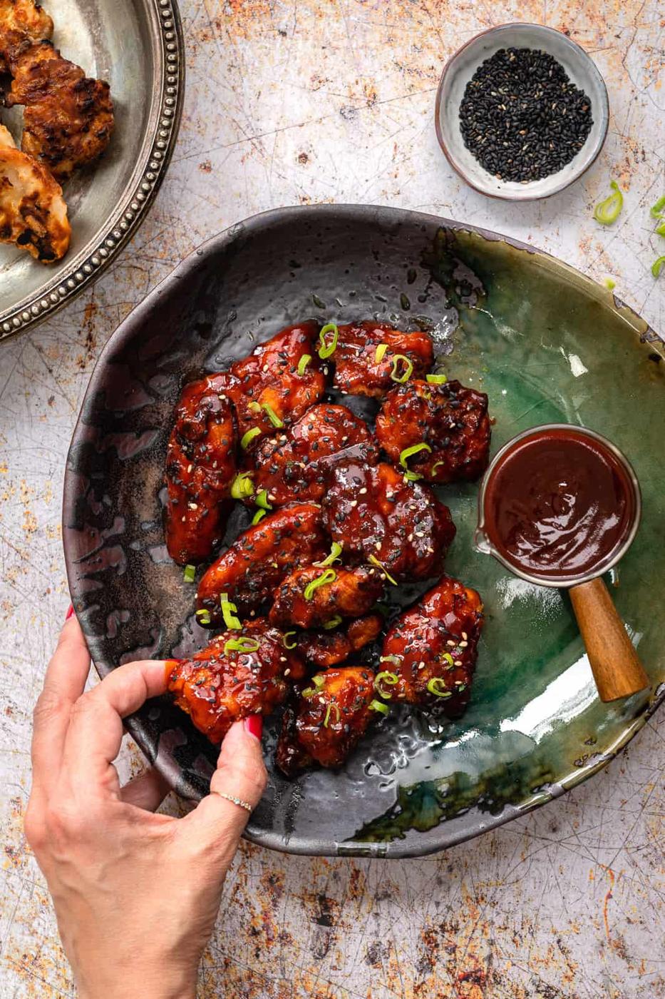 A hand reaches for a piece of saucy Korean Fried chicken wings on a green and gray ceramic plate, garnished with chopped green onions, with a small bowl of sauce and black sesame seeds nearby.