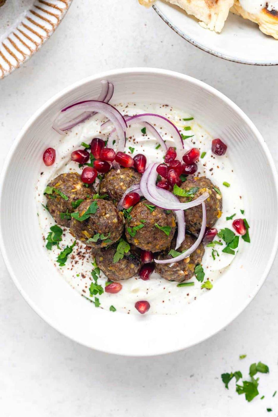 Greek meatballs served with sliced onion and pomegranate seeds over spiced yogurt in a white bowl
