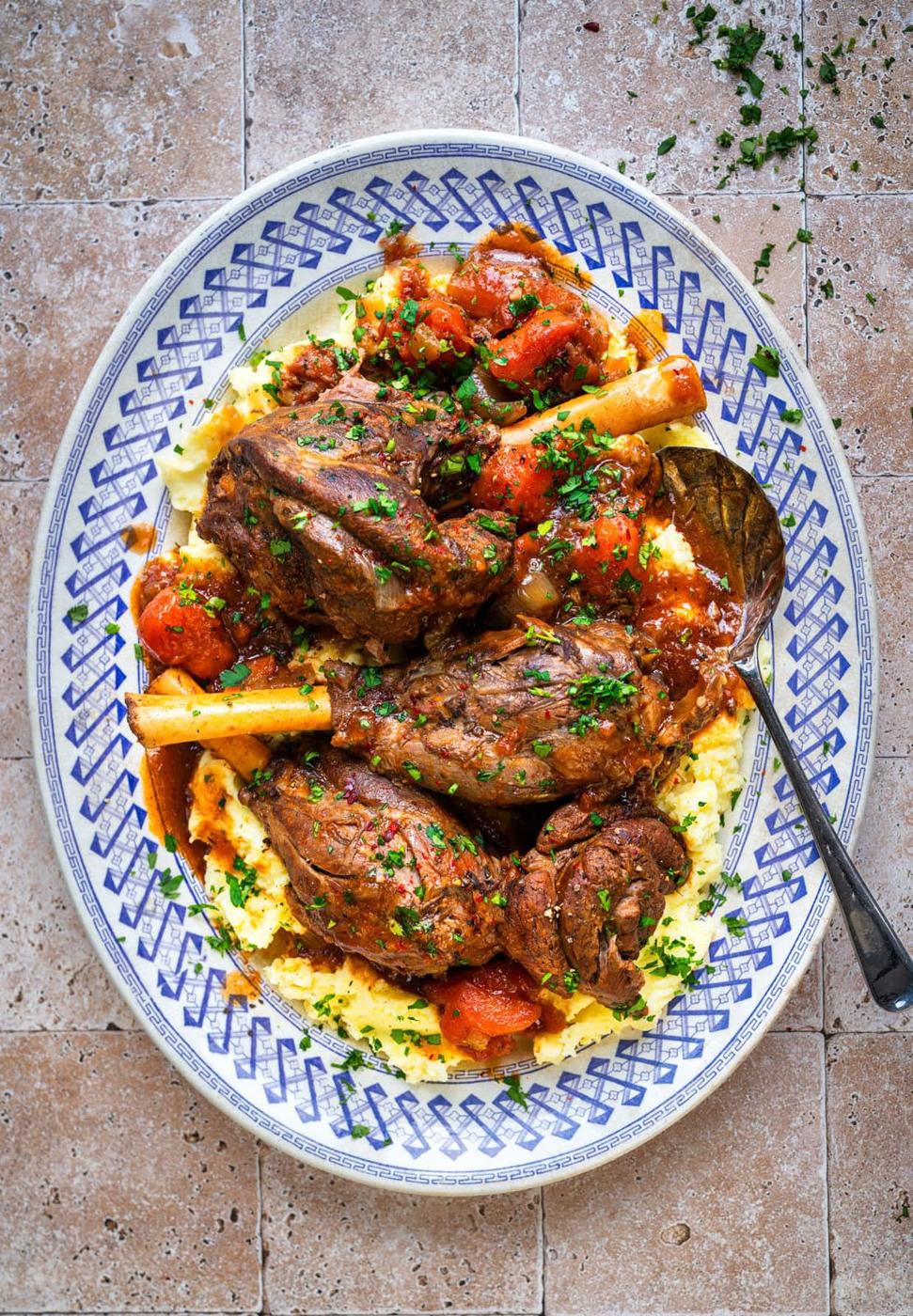 four braised lamb shanks and carrots served over mashed potatoes on a platter with red wine and tomato sauce spooned over