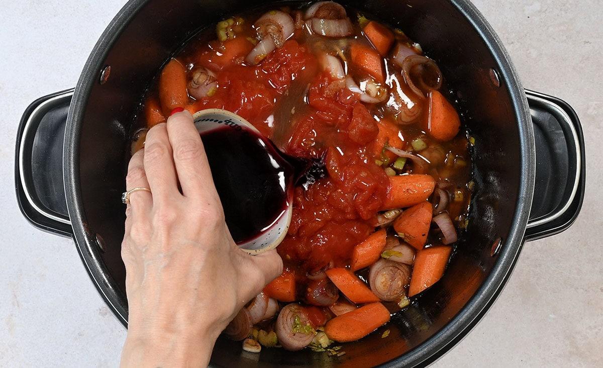adding red wine to a slow cooker with tomatoes and sautéed vegetables