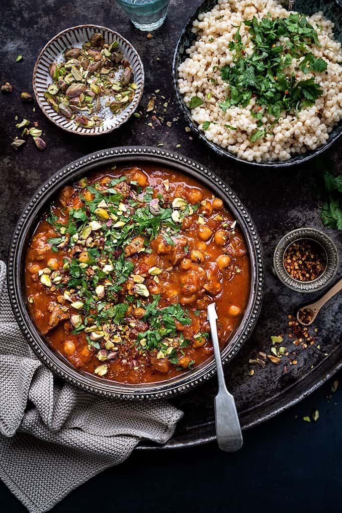 A bowl of hearty stew with chickpeas and herbs, made for comfort in the Instant Pot, is garnished with chopped pistachios, beside couscous topped with fresh herbs, a small bowl of nuts, crushed red pepper, and a gray napkin.