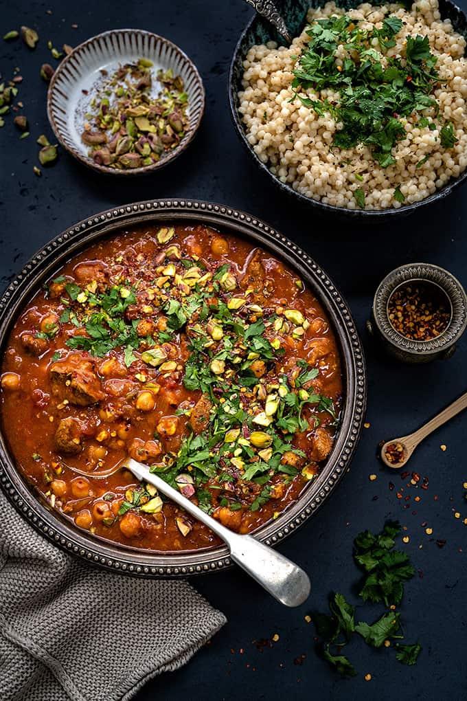 A bowl of Moroccan comfort stew with chickpeas, garnished with chopped herbs and pistachios, sits beside a bowl of couscous topped with fresh parsley, a bowl of pistachios, and a small dish of red pepper flakes.