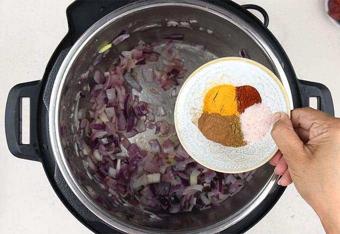 A hand holds a small bowl with Moroccan spices—turmeric, cinnamon, paprika, and salt—above an Instant Pot filled with chopped, sautéed red onions.