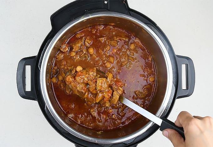 A hand stirs a pot of stew with chunks of meat and beans in a rich, red sauce inside an Instant Pot electric pressure cooker.