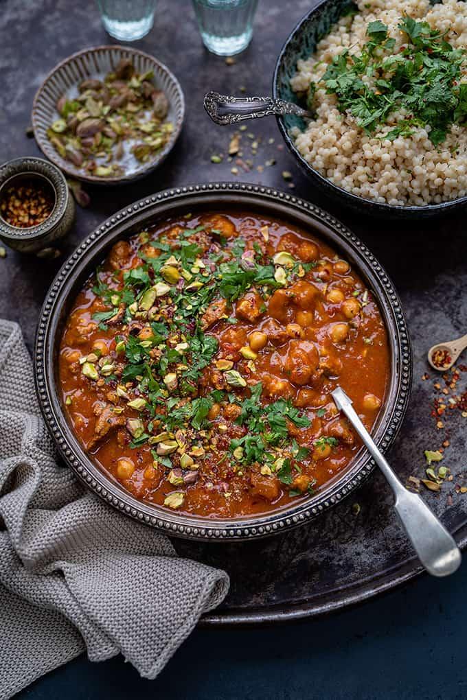 A bowl of chickpea and tomato stew, reminiscent of a vegetarian lamb tagine, garnished with chopped herbs and pistachios. Served with couscous and toppings on a dark tray, with a spoon and textured napkin beside the bowl.