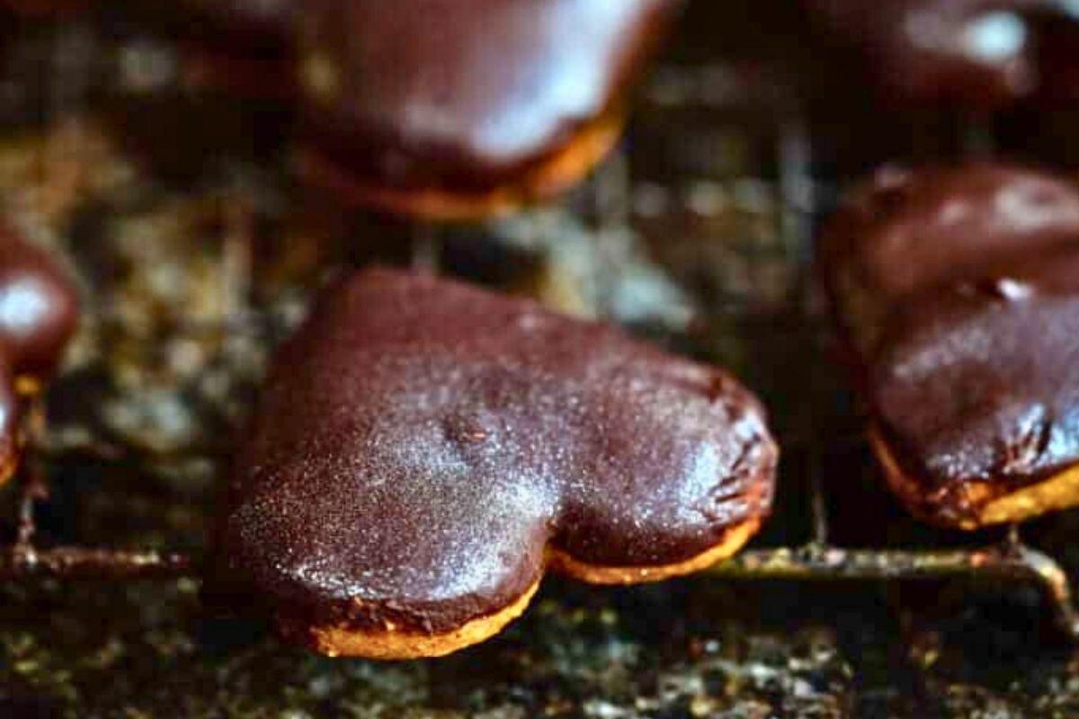 Heart-shaped lebkuchen cookies coated in chocolate glaze are cooling on a wire rack, set against a rustic brown background with a soft focus on the cookies in the back.