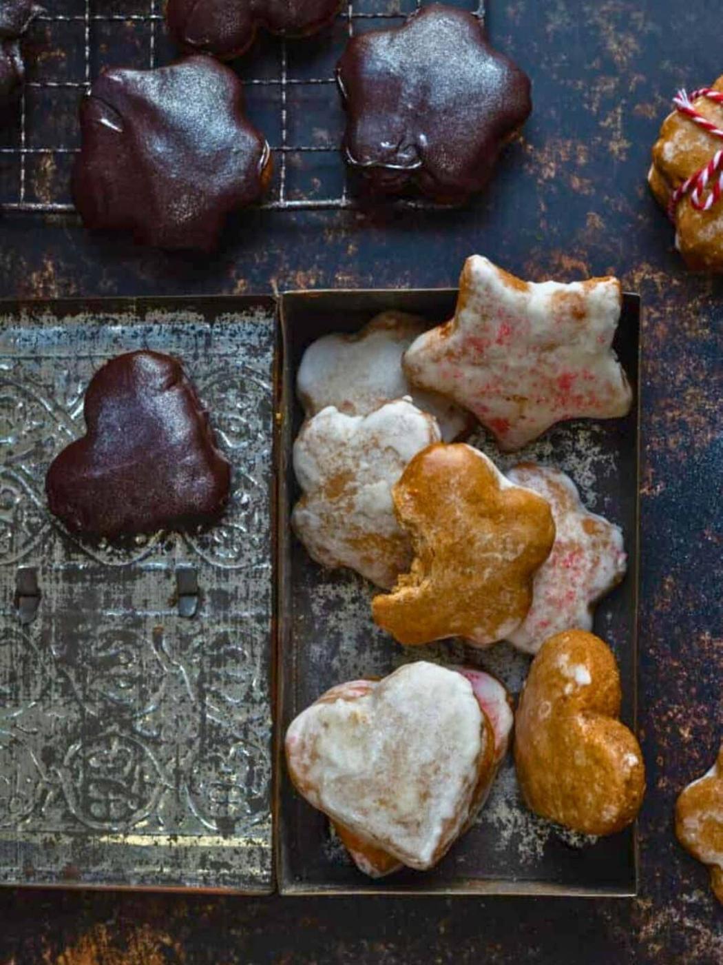 Assorted heart- and star-shaped Lebkuchen cookies with white, pink, and chocolate glazes sit in a tin and on a wire rack over a dark rustic surface. Some are partially eaten, and one shows a bite—perfect for your next lebkuchen recipe.