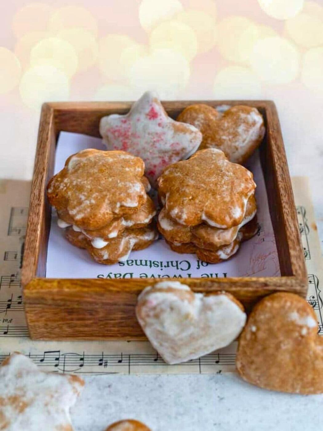 A wooden box filled with star- and flower-shaped Lebkuchen cookies, some with white glaze. A heart-shaped cookie and a partially visible cookie rest outside the box, which sits on sheet music.