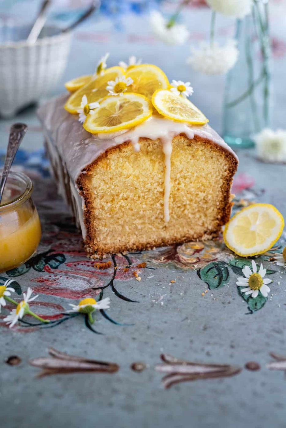 A loaf of lemon curd cake topped with icing, lemon slices, and small white flowers sits on a decorated table, with a lemon slice and a glass jar of curd nearby.