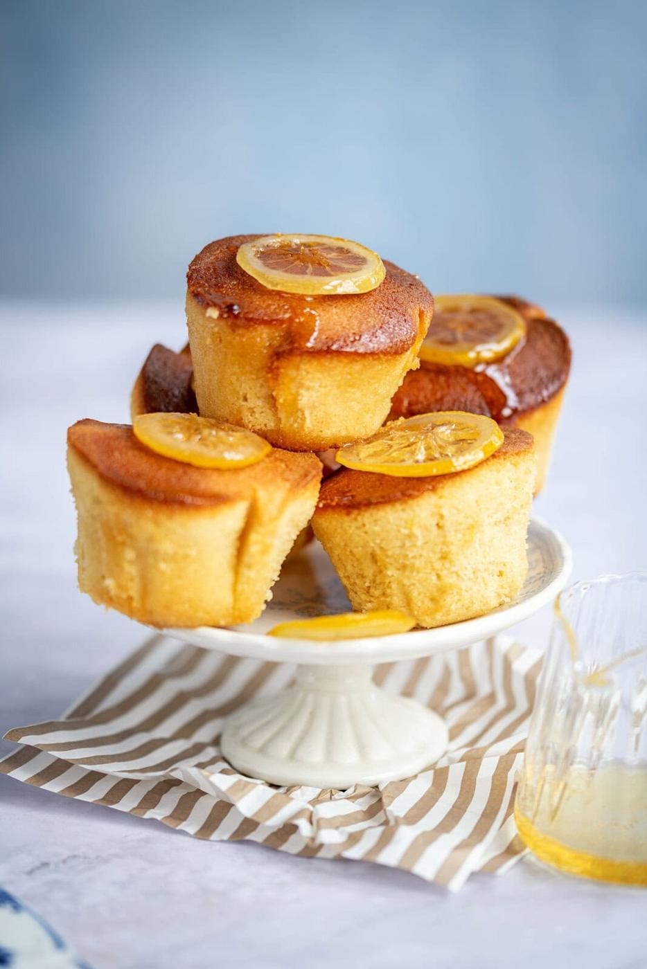 A small cake stand holds four golden lemon curd muffins, each topped with a thin slice of lemon, with a glass jar of syrup and a striped napkin nearby on a light surface.