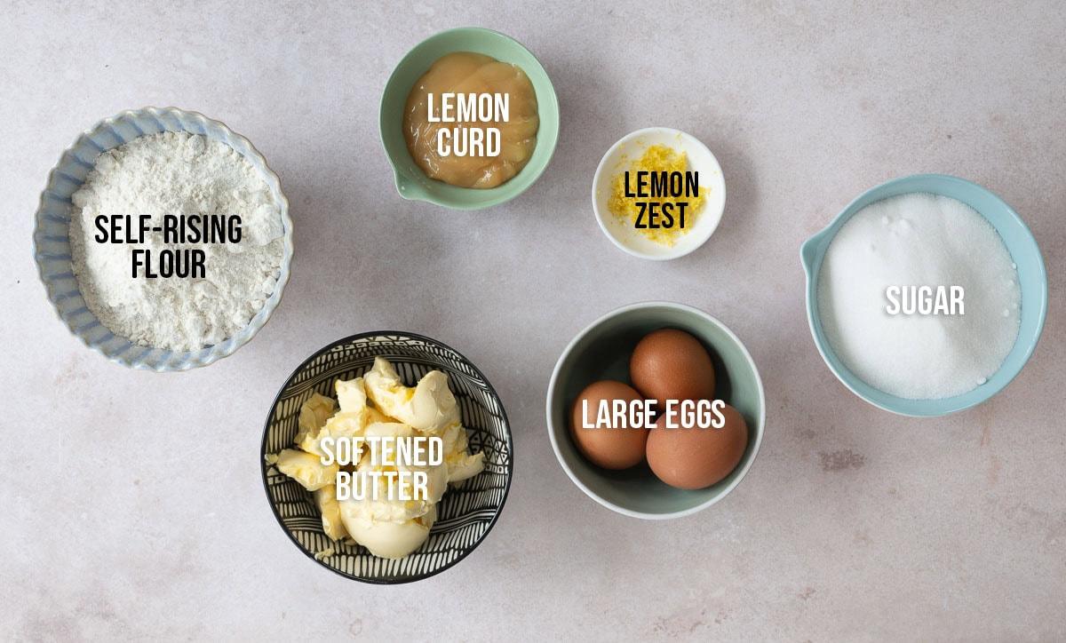 Six bowls containing labeled baking ingredients on a countertop: self-rising flour, softened butter, lemon curd, lemon zest, large eggs, and sugar.