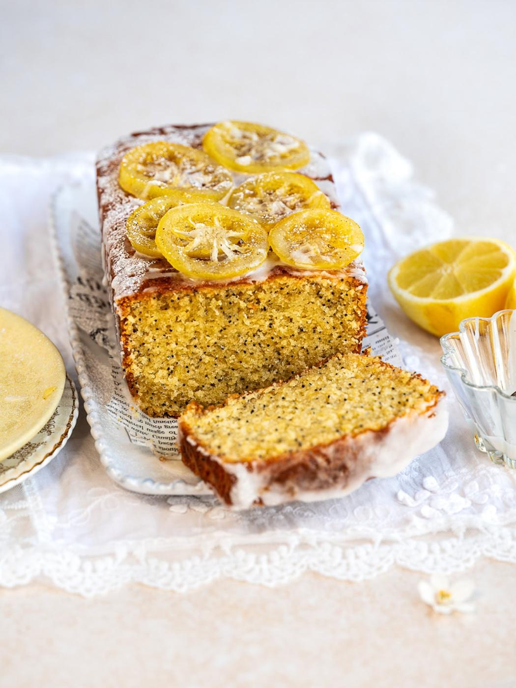 A loaf of lemon poppy seed cake topped with glazed lemon slices sits on a tray with a slice cut. A halved lemon, glass dish, and a small plate are nearby, all on a lace cloth.