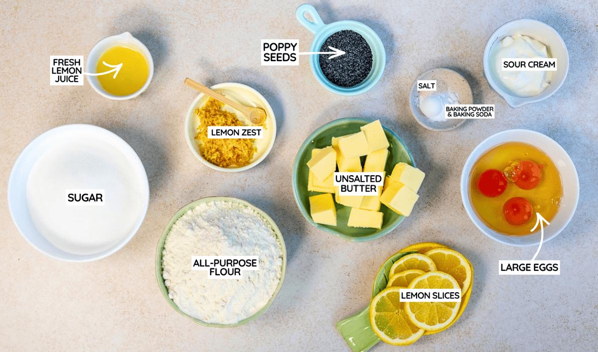 Top-down view of baking ingredients in bowls, including flour, sugar, eggs, butter, sour cream, lemon juice, lemon zest, lemon slices, poppy seeds, salt, and baking powder & baking soda, all labeled for a recipe.