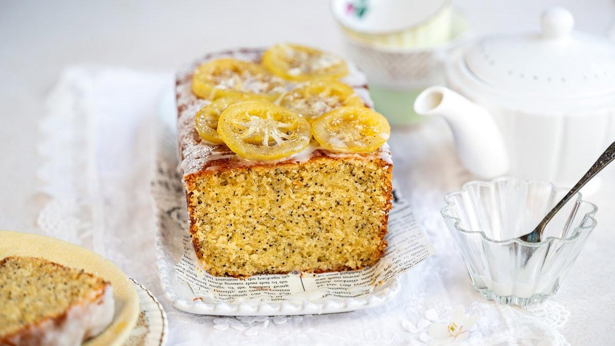 A loaf of lemon poppy seed bread topped with candied lemon slices sits on a tray lined with paper. In the background, there are teacups, a white teapot, and a glass bowl with a spoon.