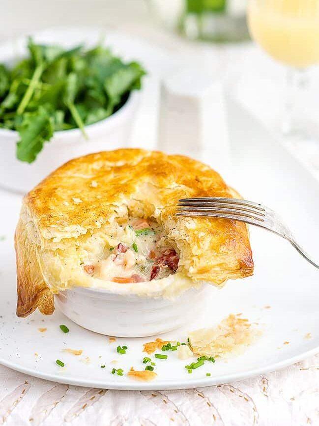A golden, flaky lobster pot pie with a piece missing reveals a creamy filling brimming with tender lobster, meat, and vegetables. A fork rests on top, while fresh greens are in a bowl in the background.