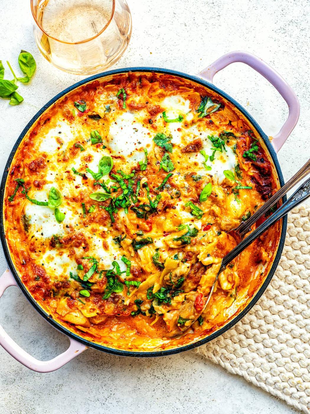 A skillet filled with cheesy baked Marry Me chicken lasagna topped with melted mozzarella and fresh basil, with serving utensils inside. A glass of white wine and a beige textured cloth are beside it on a light surface.