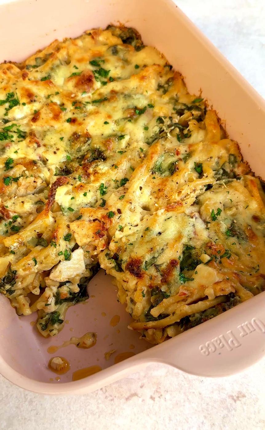 A baked casserole in a rectangular dish, featuring golden brown, melted cheese and mixed herbs on top. The dish is partially cut, revealing layers of pasta and visible greens.