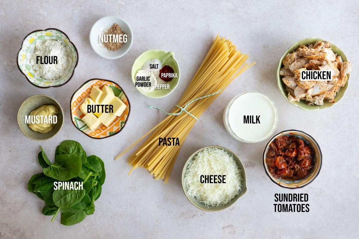 Ingredients for a pasta dish are arranged on a surface: flour, nutmeg, mustard, butter, spinach, pasta, salt, garlic powder, paprika, pepper, cheese, sundried tomatoes, milk, and chicken.
