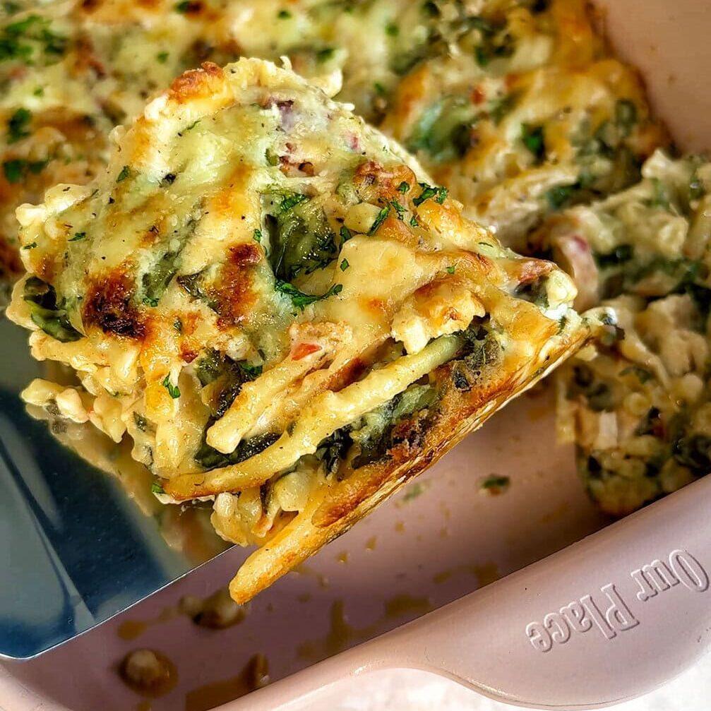 A slice of freshly baked lasagna being lifted from a baking dish. The lasagna is golden brown with melted cheese on top and visible layers of pasta, vegetables, and sauce. Sprinkled with herbs, the dish looks appetizing and savory.