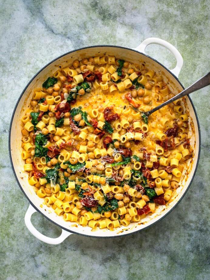 A large white pan filled with creamy pasta, sun-dried tomatoes, spinach, and Marry Me Chickpeas, topped with grated cheese, sits on a textured green surface. A metal spoon rests inside the pan.