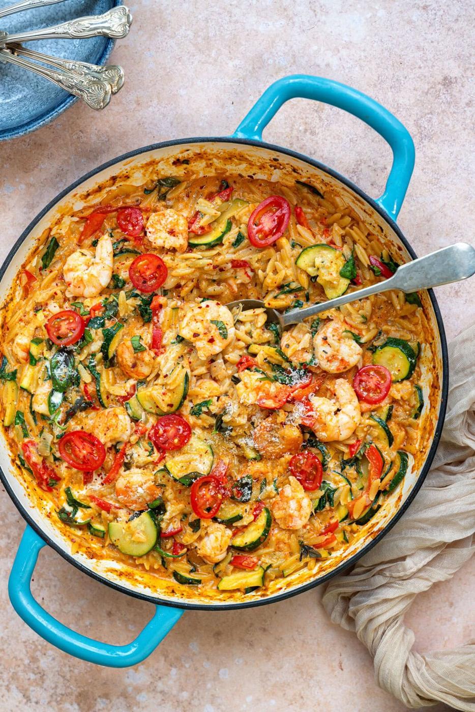 A blue-handled pan filled with creamy orzo pasta, shrimp, sliced zucchini, cherry tomatoes, and spinach, garnished with herbs and parmesan. A serving spoon rests inside the pan. Nearby are stacked plates and a beige cloth.