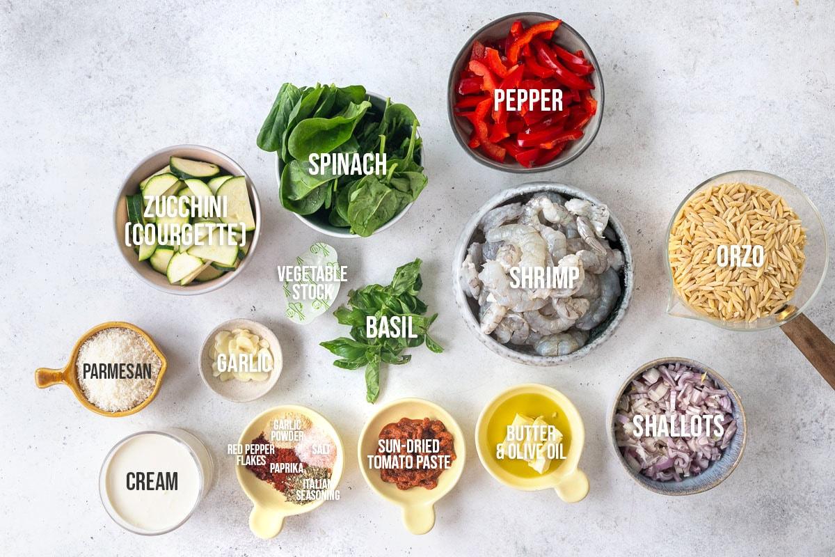 Ingredients for a shrimp orzo dish are arranged on a white surface, including zucchini, spinach, red pepper, shrimp, orzo, shallots, basil, garlic, parmesan, cream, vegetable stock, spices, sun-dried tomato paste, and butter/olive oil.
