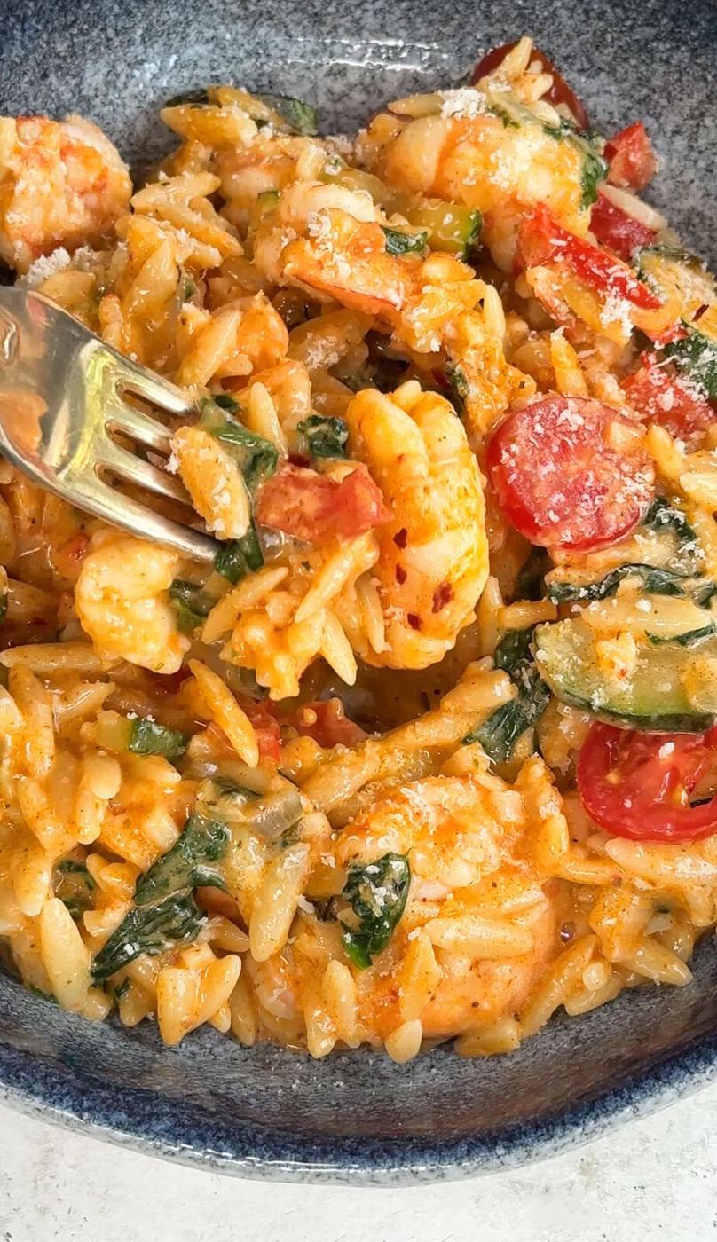 Close-up of a bowl of orzo pasta mixed with shrimp, spinach, halved cherry tomatoes, and a creamy sauce, topped with grated cheese. A fork is spearing a bite-sized portion.