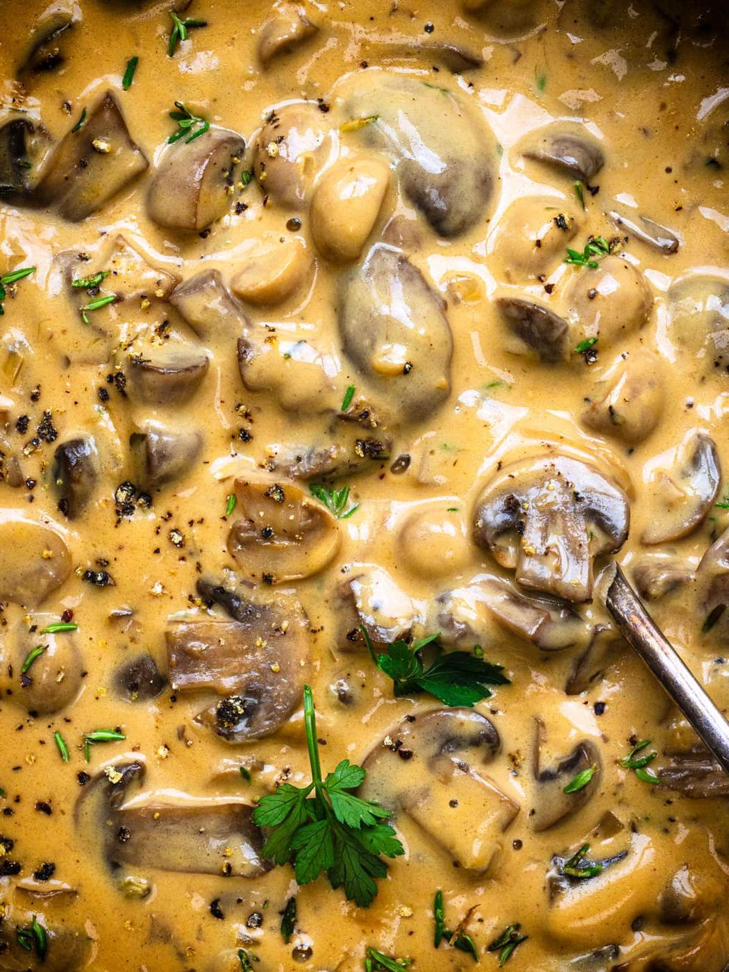 Close-up of creamy mushroom Marsala sauce with sliced mushrooms, black pepper, and fresh parsley, with a spoon partially visible, garnished with herbs.