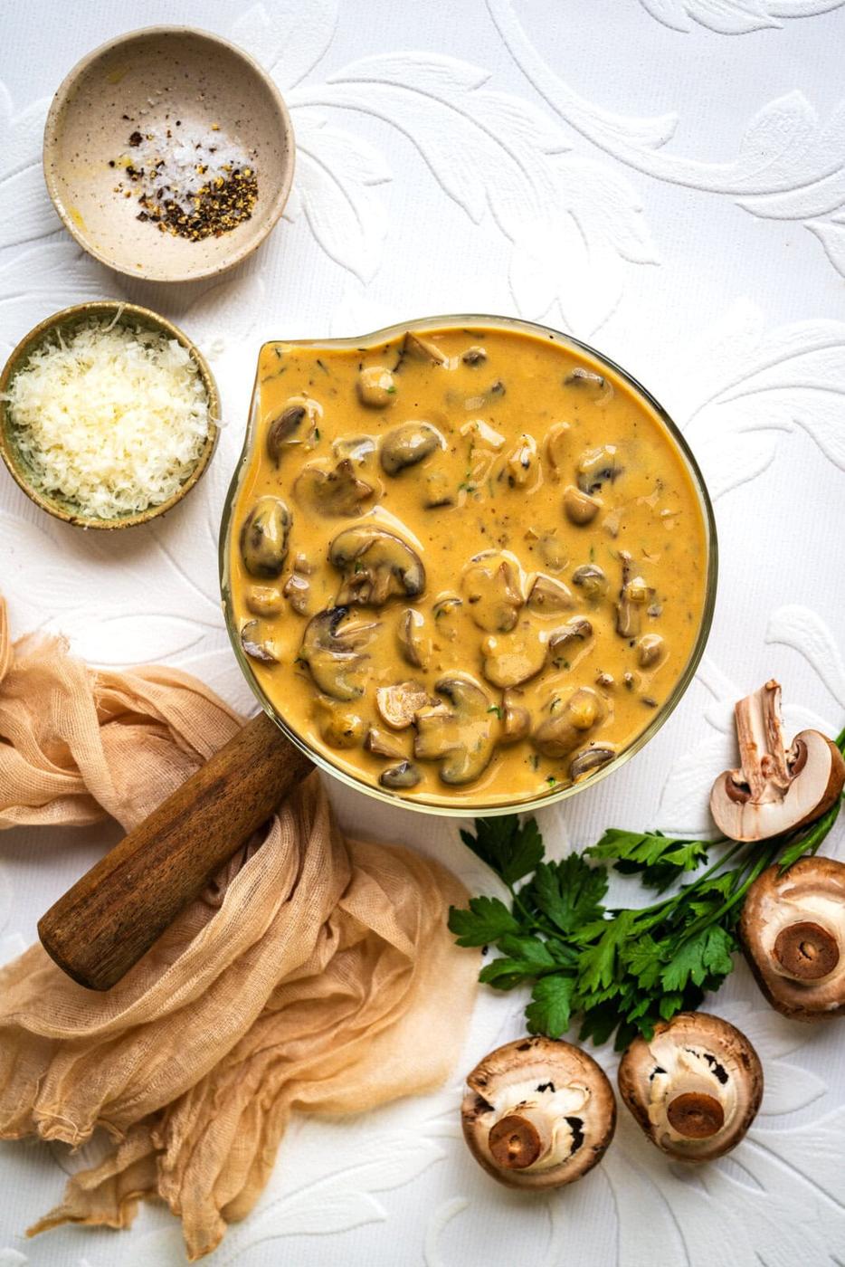 A bowl of creamy Marsala sauce with sliced mushrooms, surrounded by fresh mushrooms, parsley, grated cheese, a small bowl of pepper, and a wooden rolling pin on a textured white surface.