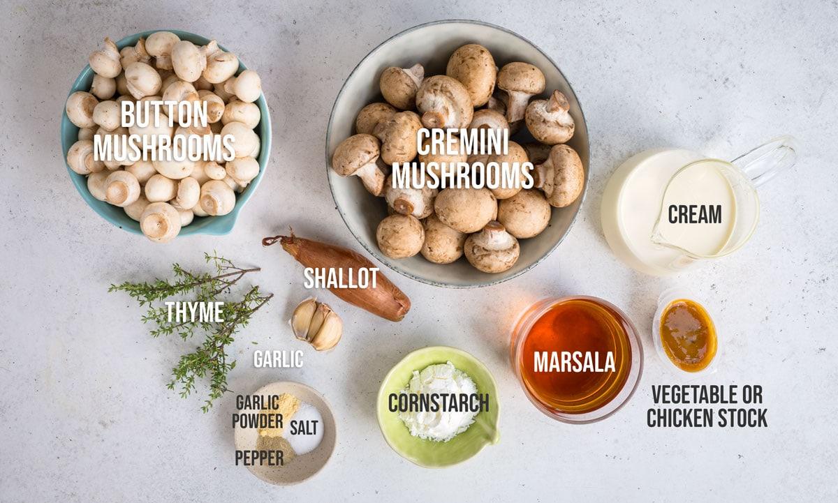 A top view of labeled ingredients for a recipe of mushrom marsala sauce, including button mushrooms, cremini mushrooms, cream, shallot, thyme, garlic, marsala, cornstarch, vegetable or chicken stock, garlic powder, salt, and pepper.