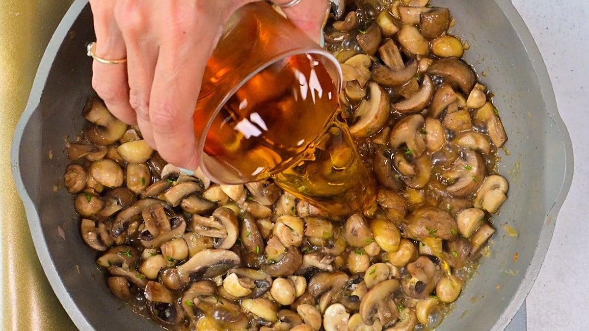 A hand pours a glass of Marsala wine into a pan filled with sautéed mushrooms, sliced and whole, on a stovetop.