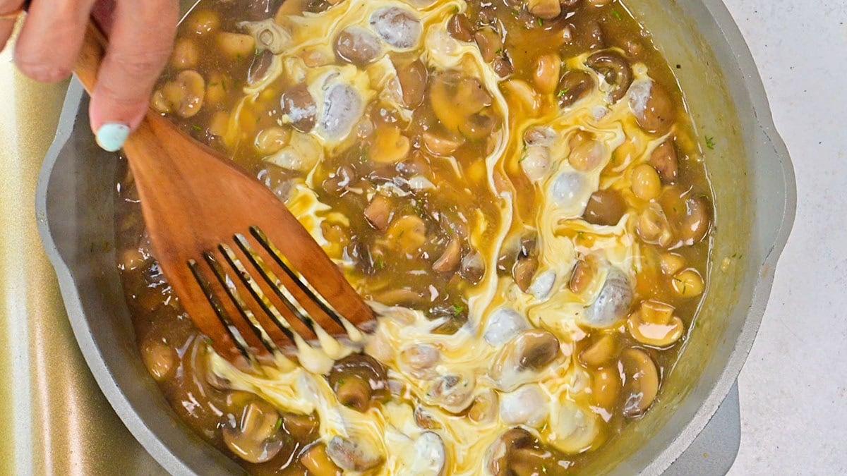 A hand with light blue nail polish stirs a creamy mushroom sauce with a wooden spatula in a light-colored pan. The mixture has visible mushrooms and swirling cream throughout.