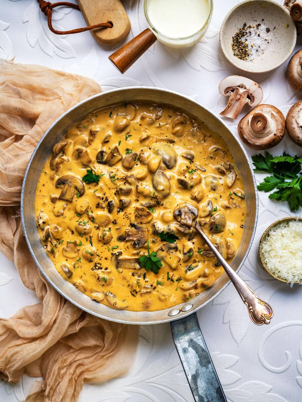 A skillet filled with creamy mushroom Marsala sauce garnished with parsley, surrounded by fresh mushrooms, grated cheese, a bowl of pepper, and a glass of cream on a white textured surface.