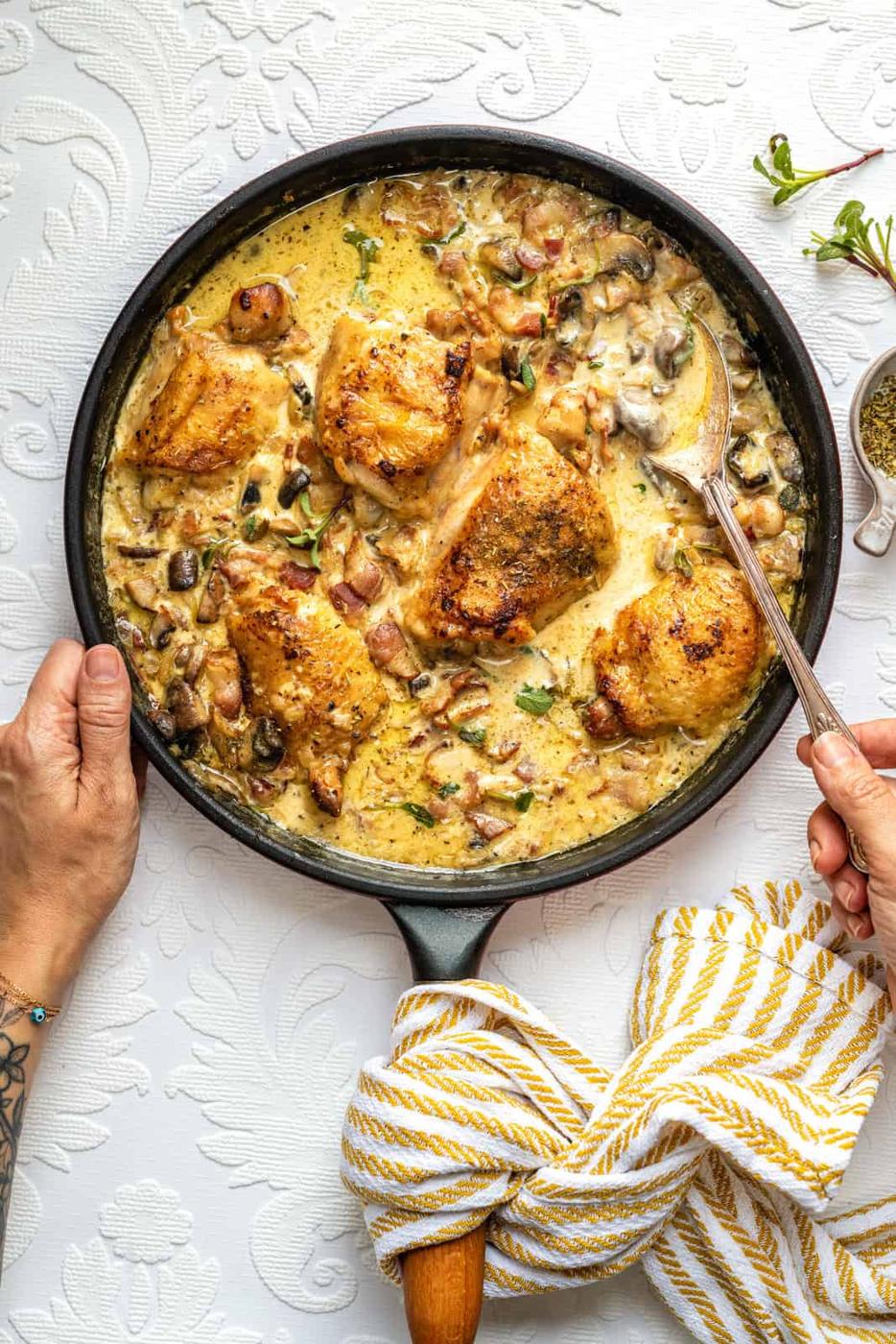 A skillet of creamy mushroom chicken with golden-brown chicken thighs, mushrooms, and herbs in a creamy sauce—reminiscent of a Mary Berry Chicken Casserole. A yellow-striped towel wraps the handle as hands serve the dish.