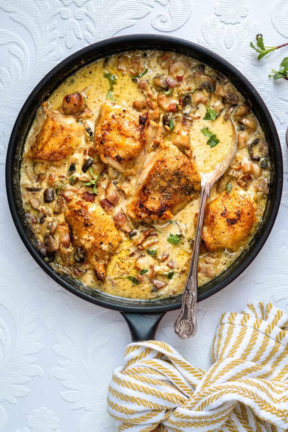A skillet filled with creamy chicken and vegetables, garnished with herbs, evokes a classic Mary Berry chicken casserole. Four browned chicken pieces rest in a thick sauce with mushrooms and diced vegetables. A spoon is in the pan, and a striped towel sits nearby.