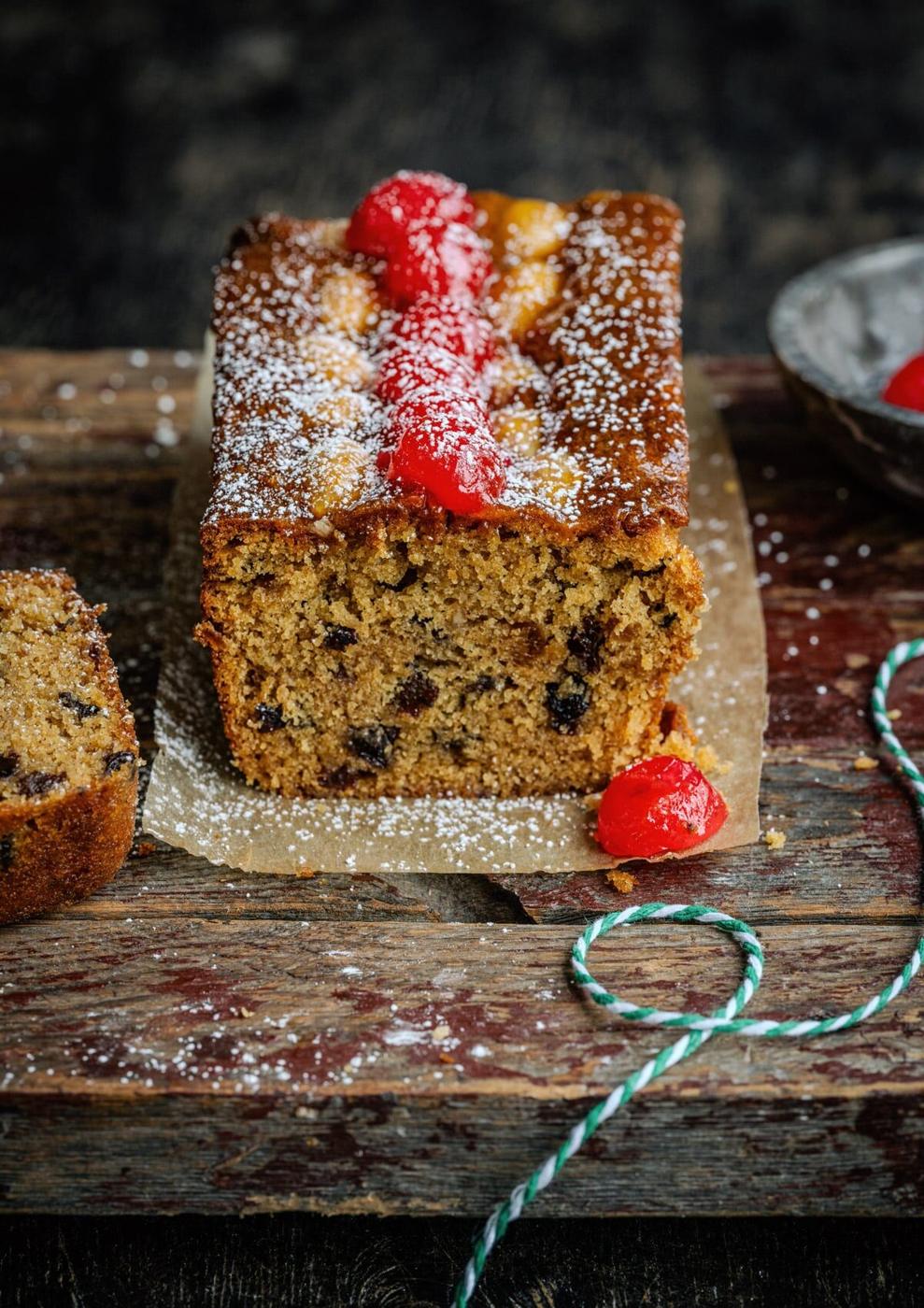 A Mincemeat Loaf Cake topped with powdered sugar and bright red cherries sits on parchment paper on a rustic wooden board, with a slice cut from one end.