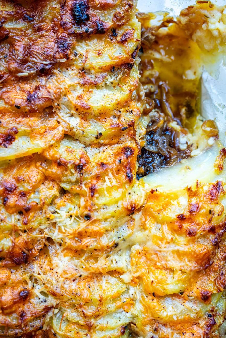 Close-up of a golden, crispy potato gratin topped with melted cheese. Thinly sliced potatoes are layered over a creamy, cheesy filling with visible browned bits and a portion scooped out to reveal the inside.