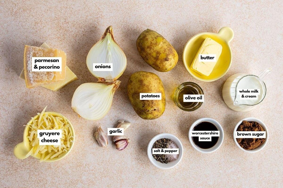 Top-down view of ingredients for a recipe, including parmesan & pecorino, onions, potatoes, butter, olive oil, whole milk & cream, gruyere cheese, garlic, salt & pepper, Worcestershire sauce, and brown sugar.