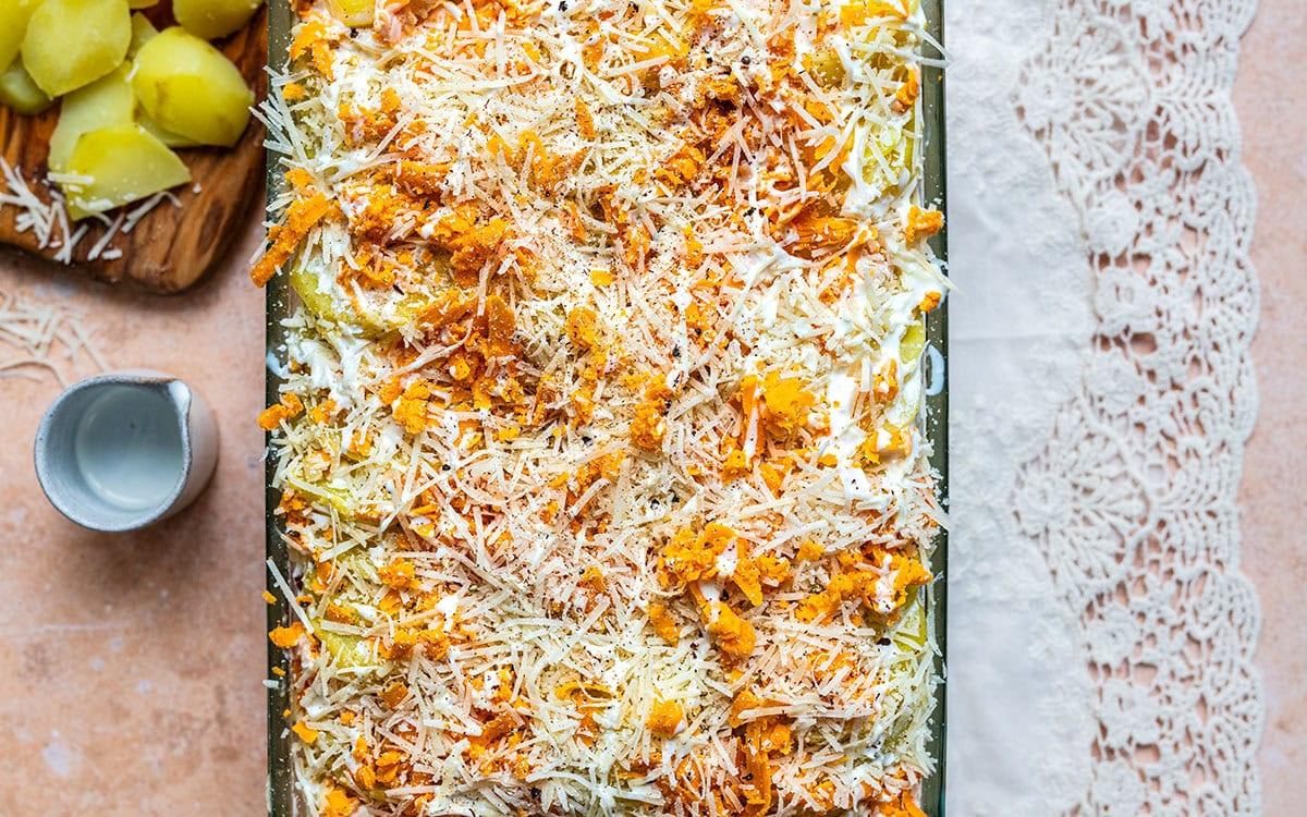 A baking dish filled with a layered casserole topped with shredded cheese and orange bits, placed on a pink surface next to boiled potato pieces on a wooden board and a cup, with a lace cloth on the right side.