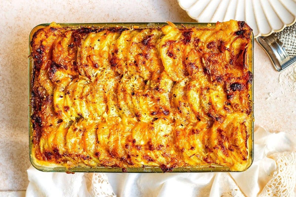A golden-brown, baked mashed potato gratin in a rectangular glass dish, featuring layers of thinly sliced potatoes, caramelized onions, and crispy potatoes topped with cheese with crisped edges. A cheese grater and white napkin sit nearby on a light surface.