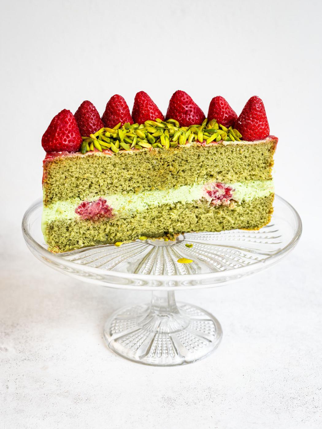 A slice of green sponge cake with a creamy filling, raspberries inside, topped with sliced pistachios and whole strawberries, displayed on a glass cake stand against a white background.