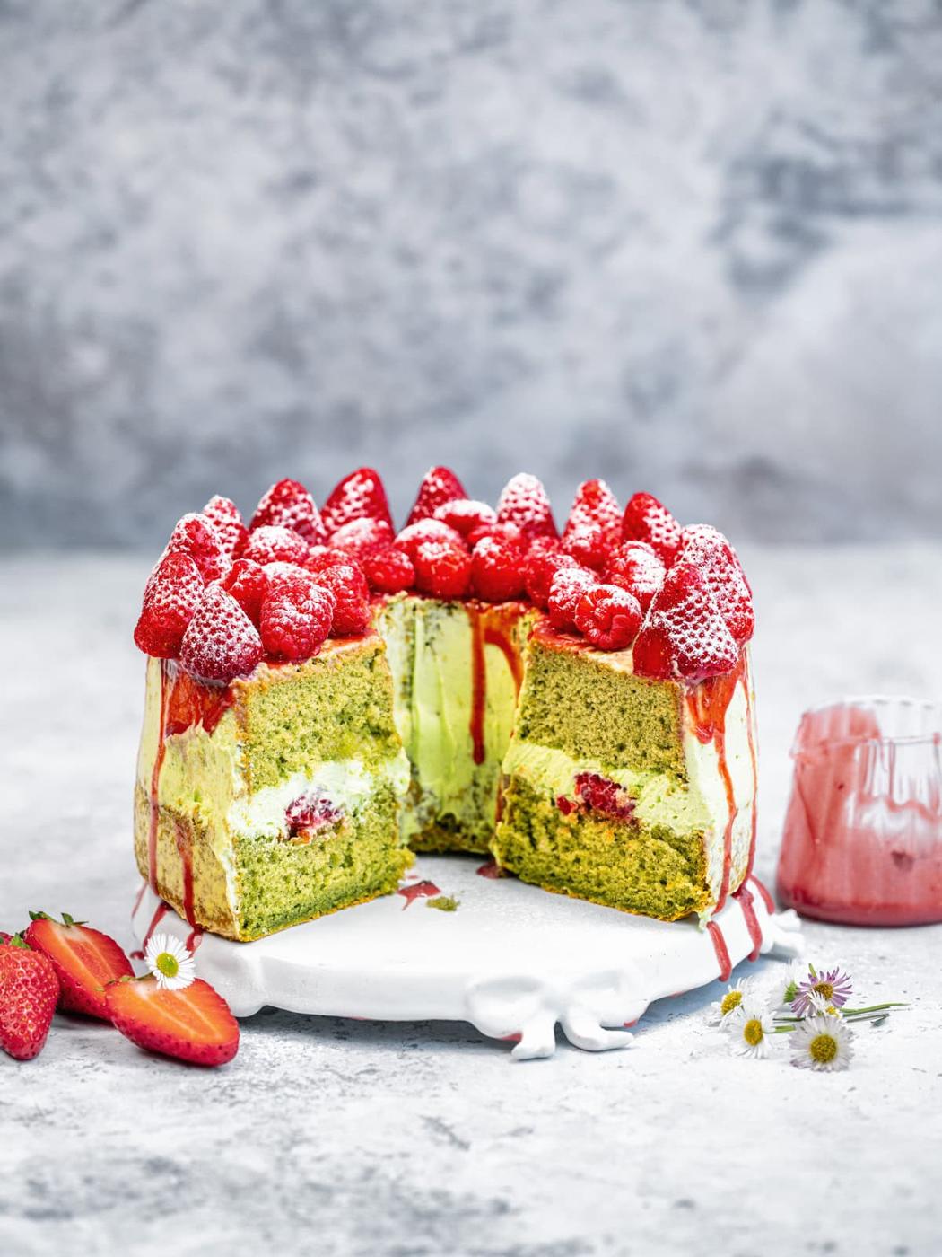 A tall matcha cake with a slice cut out, filled with cream and strawberries, topped with whole strawberries and powdered sugar, sits on a white stand; strawberries and flowers are scattered nearby.
