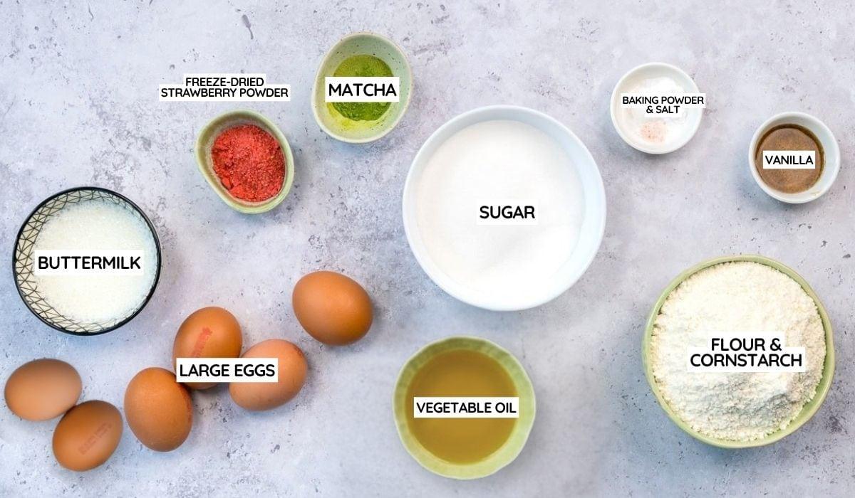 Top-down view of Matcha Cake ingredients labeled on a light surface for a Matcha Chiffon Cake Recipe: buttermilk, large eggs, freeze-dried strawberry powder, matcha, sugar, vegetable oil, flour & cornstarch, baking powder & salt, and vanilla.