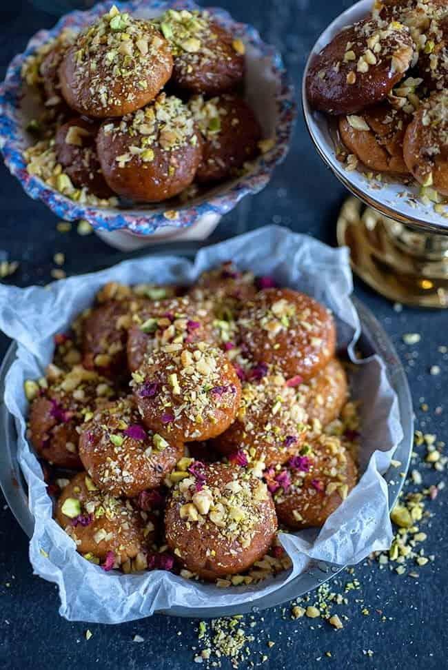 Bowls and trays filled with golden brown Melomakarona, traditional Greek Christmas Honey Cookies, are topped with chopped nuts and rose petals on a dark surface. The glossy treats are garnished with generous sprinkles of pistachios.