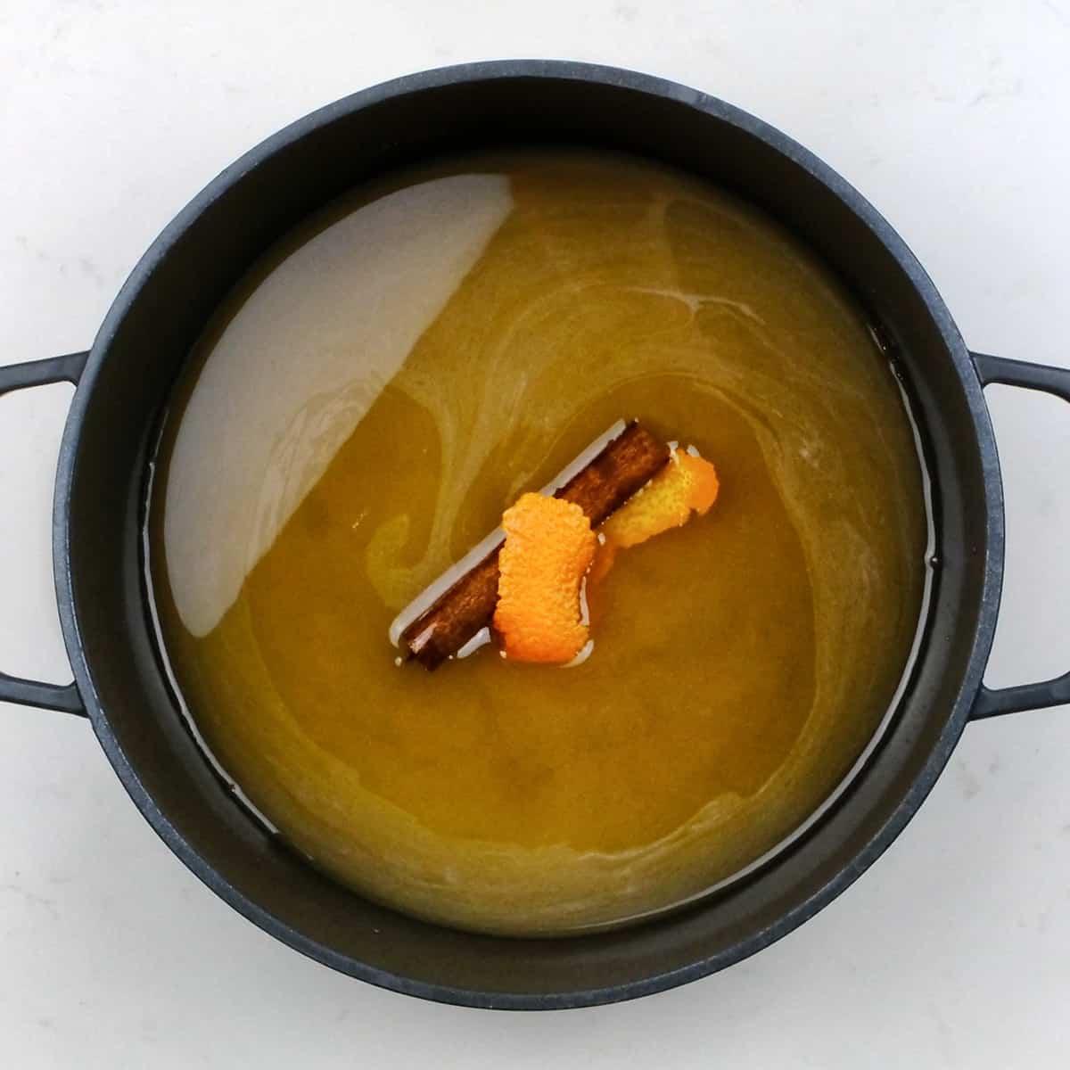A black pot contains a light brown liquid with a cinnamon stick and two orange peel strips floating on top—perfect for soaking Melomakarona, the classic Greek Christmas Honey Cookies. Seen from above on a white surface.