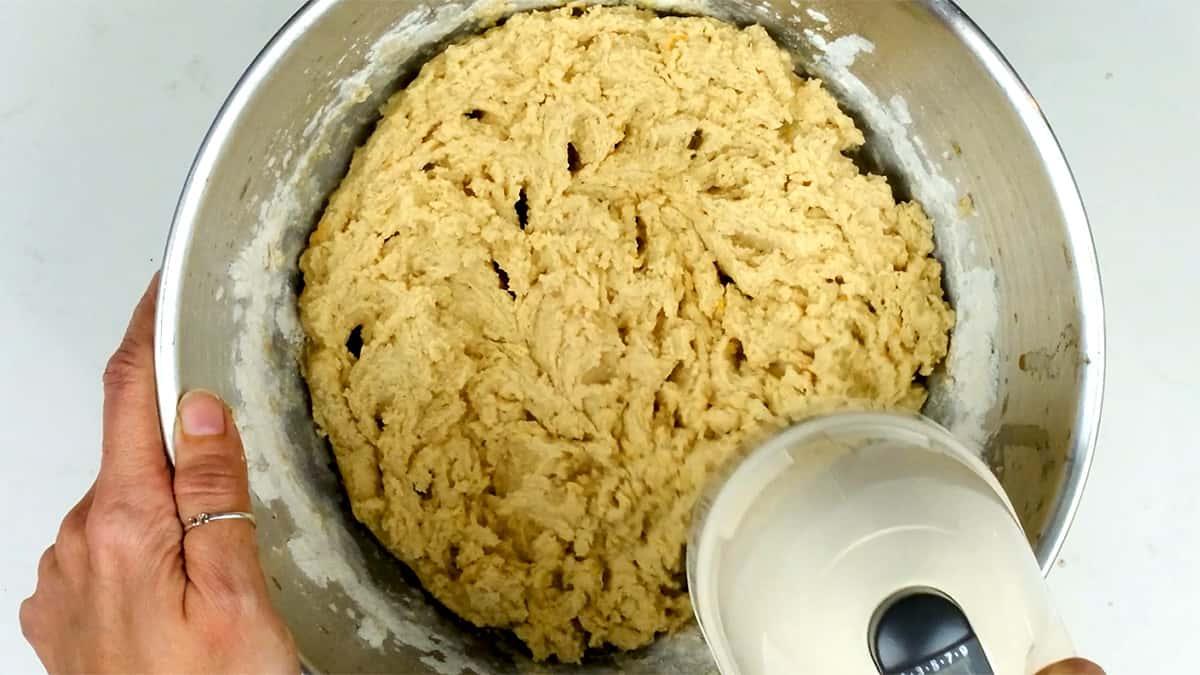 A person holds a metal bowl of thick bread dough with one hand while pouring liquid from a measuring cup into the dough with the other, preparing it for delicious Melomakarona, traditional Greek Christmas honey cookies.