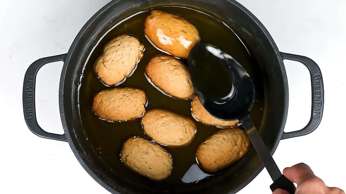 A hand uses a black spoon to baste oblong Melomakarona dough pieces frying in hot oil inside a black pot, viewed from above.